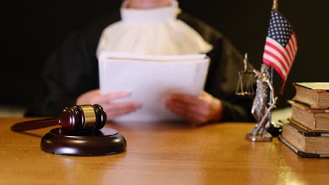 a judge is carefully examining legal papers and documents at a courtroom table, with a gavel and american flag present