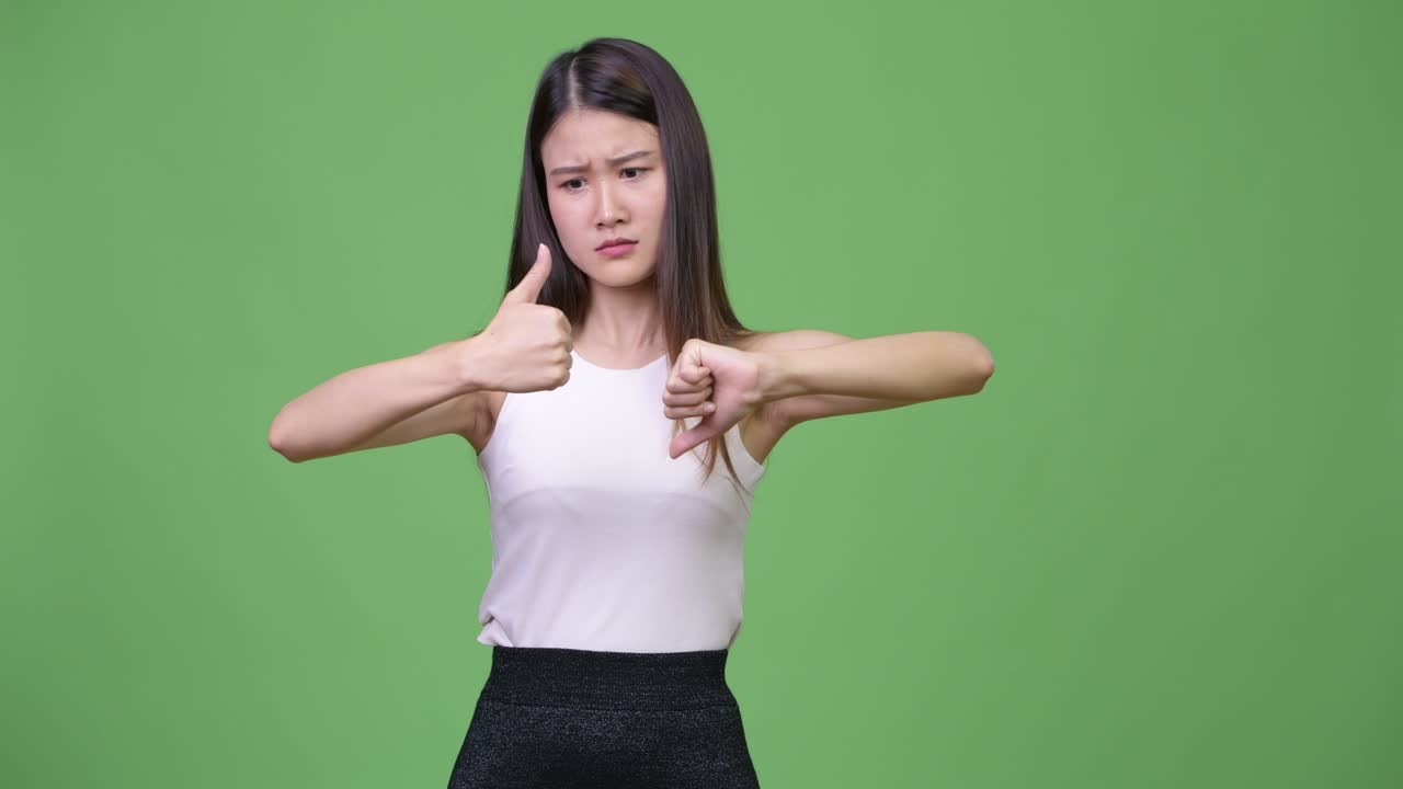 Young confused Asian businesswoman choosing between thumbs up and thumbs down