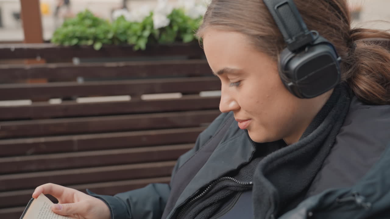 Giovane donna che legge un libro su una terrazza, avvolta in una giacca e con le cuffie. Gira lentamente le pagine, tazza di caffè caldo su un tavolino di vetro, balcone urbano con piante, primi piani spontanei che catturano una concentrazione pacifica
