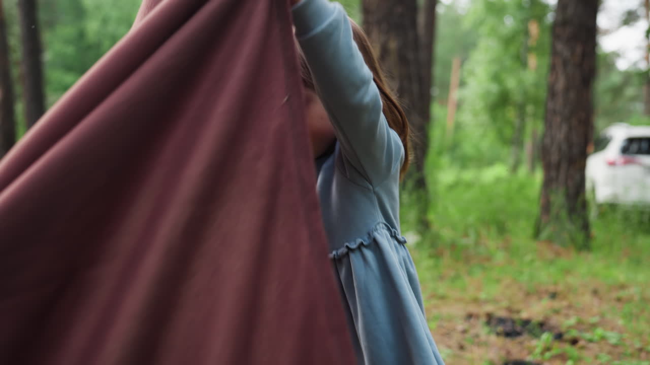 Niño pequeño sosteniendo el extremo de una tela marrón mientras la muerde y la extiende en el suelo del bosque con un coche aparcado al fondo, mostrando expresión natural, concentración y un momento lúdico al aire libre