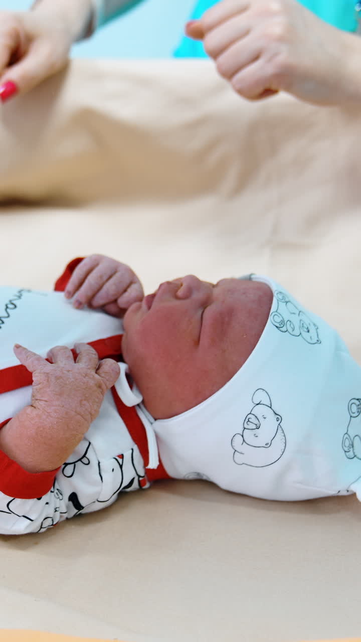 Tiny little child in a cap and sleepsuit lies on the table laid with sheets. Nurse in latex gloves puts on warm sock on baby's feet. Vertical video