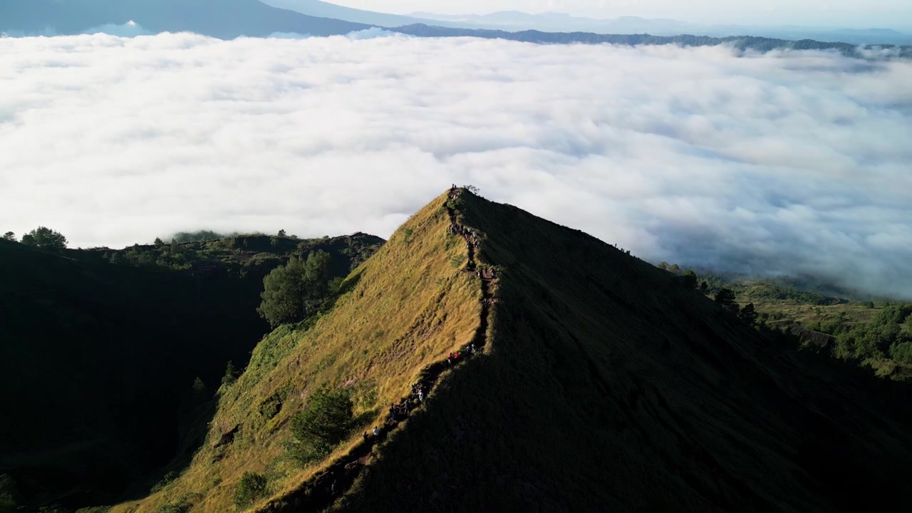 Trilha de cume de montanha com pessoas caminhando acima de um mar de nuvens sob a luz quente da manhã, através da paisagem vulcânica de Bali, com terreno verde acidentado e vales profundos, em um recuo de drone com revelação em inclinação ascendente.