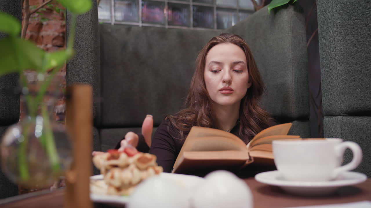 Lady dressed in black sits comfortably inside modern booth reading brown book, gently flipping page while using finger to scoop cream from plate beside waffles and coffee on wooden table