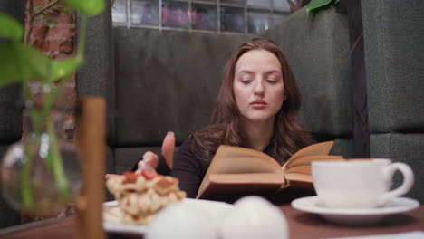 lady dressed in black sits comfortably inside modern booth reading brown book, gently flipping page while using finger to scoop cream from plate beside waffles and coffee on wooden table