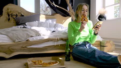 young blonde woman using mobile phone and eating pizza while sitting on the floor in the bedroom at home