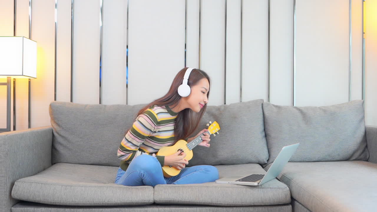 귀여운 젊은 여성이 소파에 앉아 노트북과 상호 작용하면서 노란색 우쿨렐레를 연주하는 것을 즐깁니다.