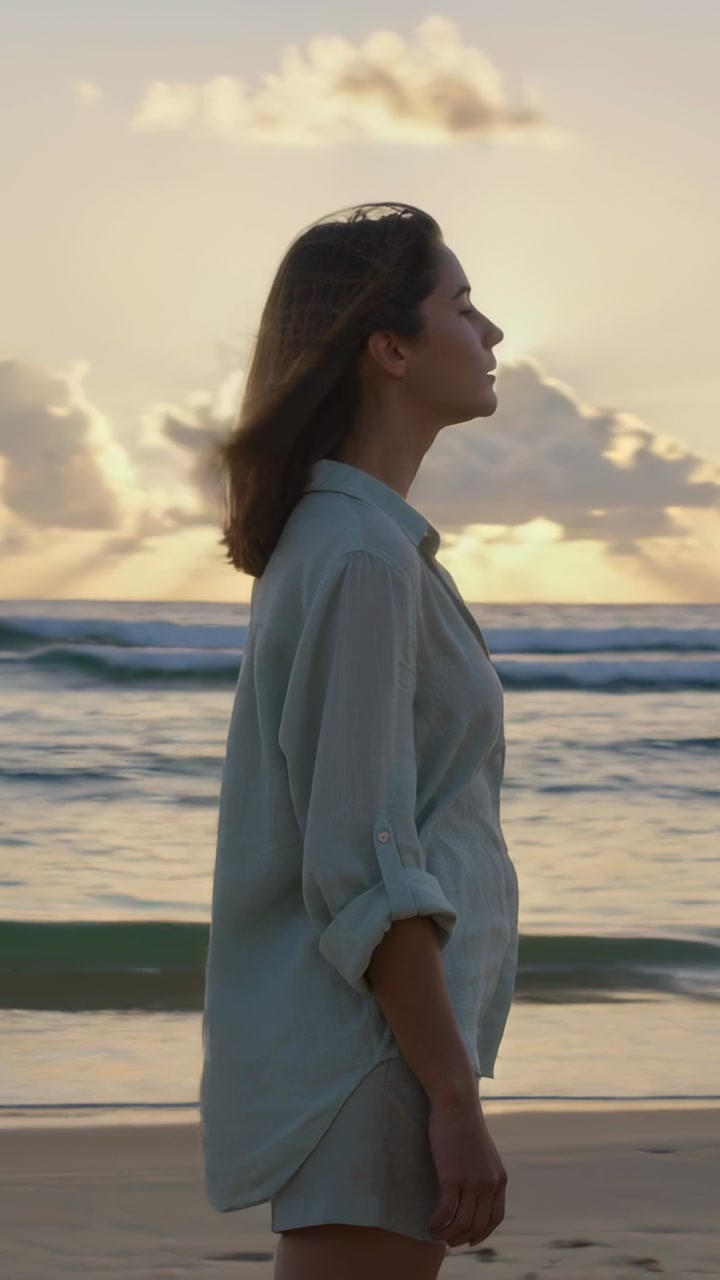 Vertical video: Walking woman in loose shirt and shorts along shore at sunrise, breeze lifting hair