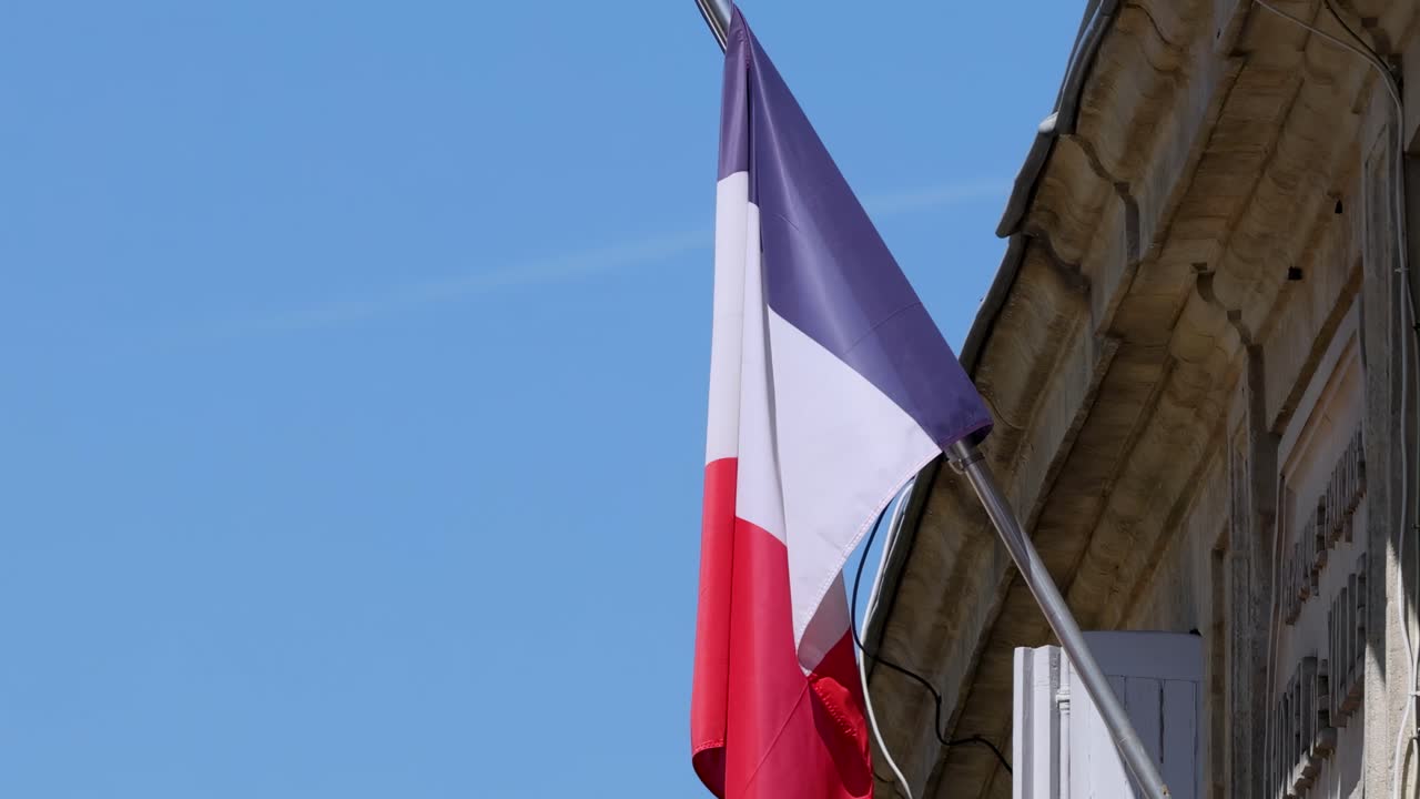 A close-up of the French flag waving on a historic stone building under a clear blue sky.