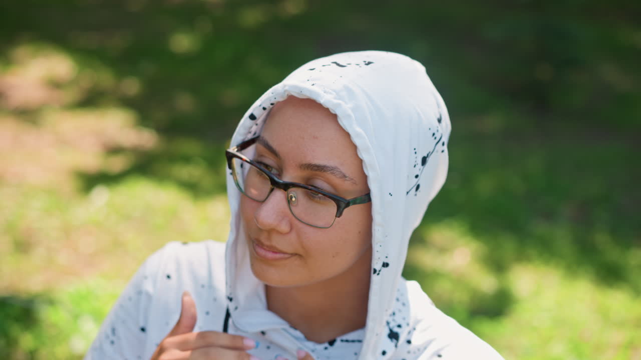 햇빛이 잘 드는 공원에서 후드티를 벗고 있는 흑인 여성의 야외 클로즈업 사진, 안경, 땋은 머리를 뒤로 묶음, 생각에 잠긴 표정, 학생의 몸짓, 통근자의 잠시 멈춤, 부드러운 보케 배경, 자연광