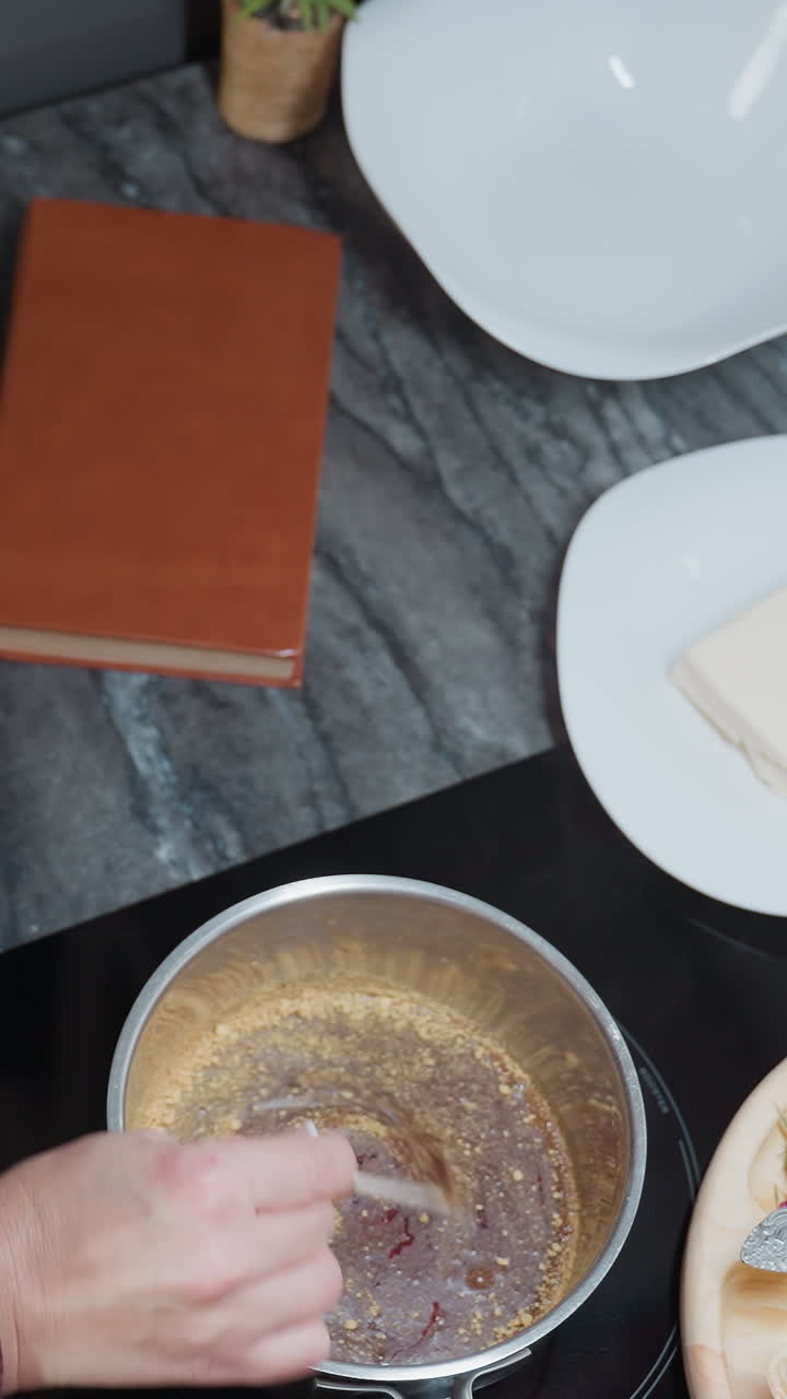 vista de arriba hacia abajo de una mujer agitando sopa en una olla de acero inoxidable usando un palo, con varias herramientas de cocina, ingredientes, bandeja de madera e artículos de cocina dispuestos en una moderna encimera de mármol oscuro