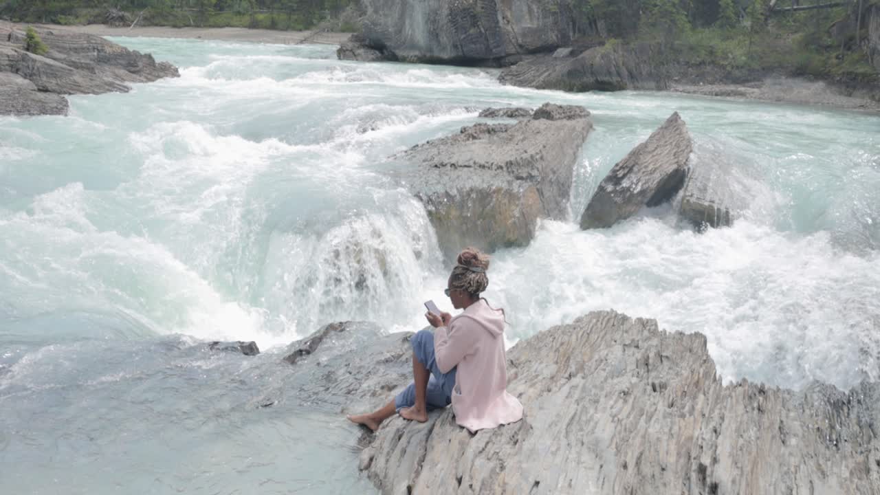 Black lady on her phone sitting on the rocks near the edge of a waterfall (Natural Bridge lower falls) and beautiful scenic blue river in Yoho National Park