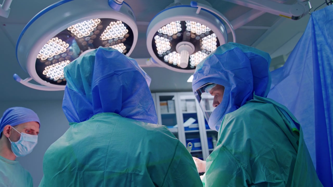 Équipe chirurgica che collabora ed esegue un'operazione ortopedica. Medici con caschi protettivi lavorano sotto le luminose e moderne lampade rotonde.