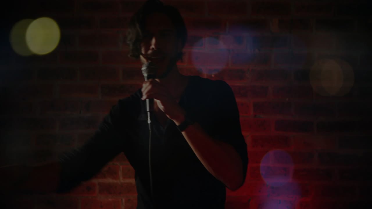 Un comico si avvicina al microfono su indicazione delle luci, eseguendo battute per far ridere mentre i bagliori bokeh gli mascherano il volto.