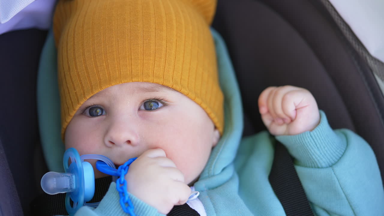 Healthy Caucasian baby boy in warm clothes lies in stroller during walk outside. Tiny kid looks up holding a pacifier at his mouth. Close up.