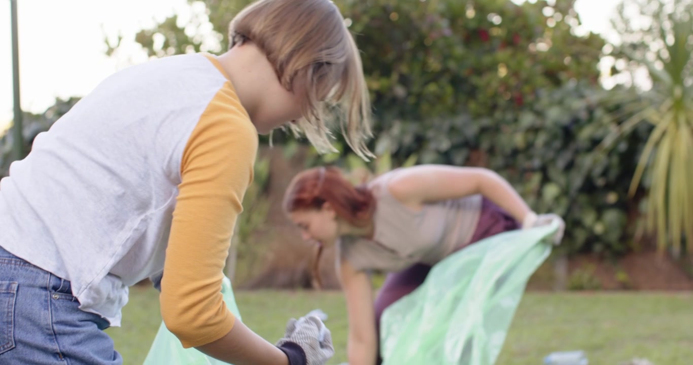 Diverse female volunteers unfolding green bag, picking up bottles and cleaning yard while gloved