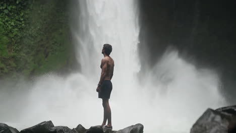 man in front of roaring waterfall in rain forest turns to look up at cascade