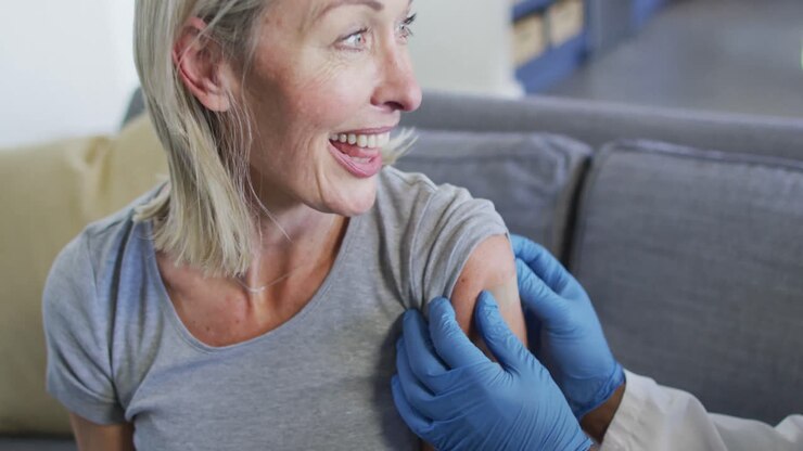 Happy senior diverse woman and doctor in living room sitting on sofa, vaccinating