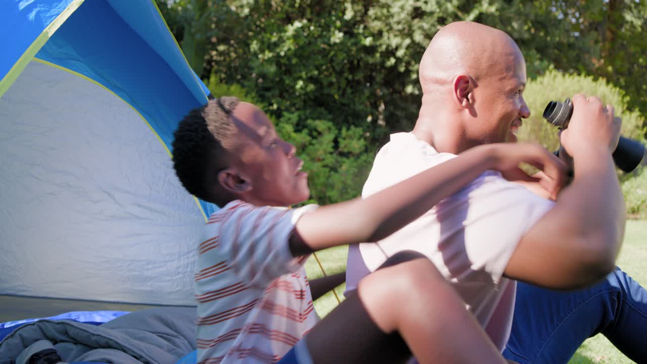 Afroamerikansk far og søn sidder på græsset ved teltet, far peger og guider sønnen, der bruger en kikkert.