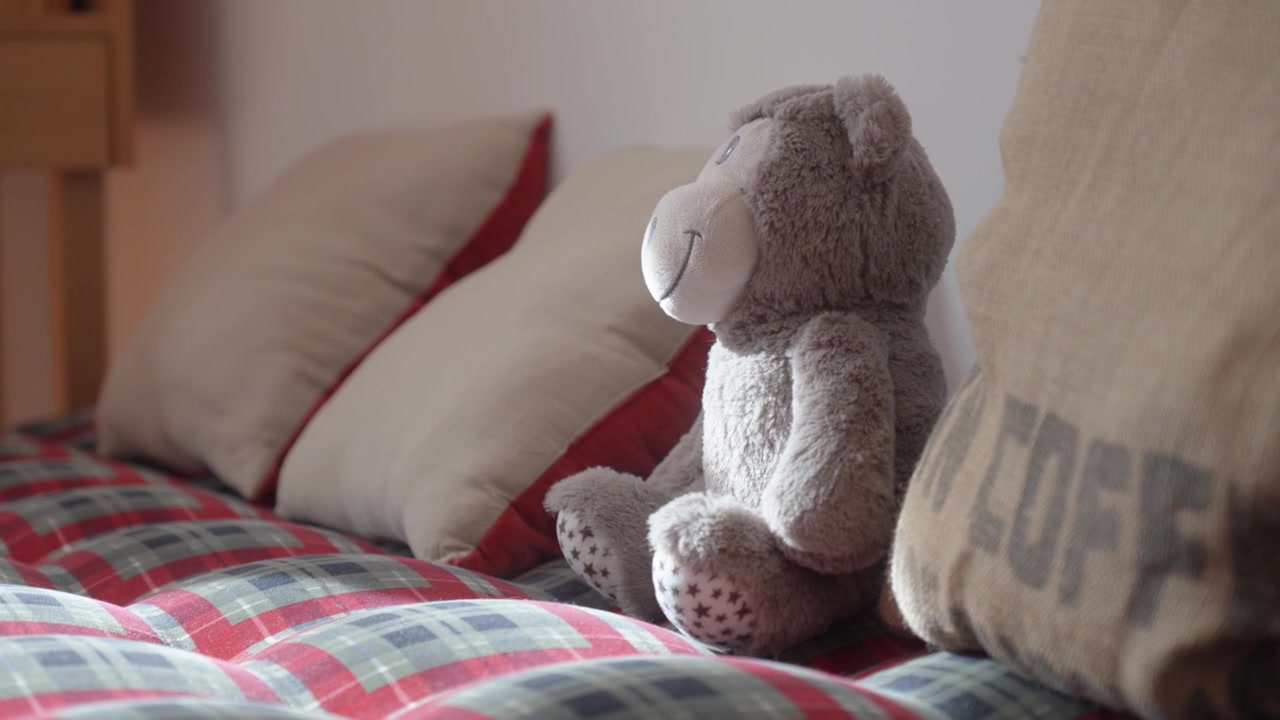 Oso de peluche sentado en la cama con almohadas decorativas en un cálido ambiente interior de hogar.