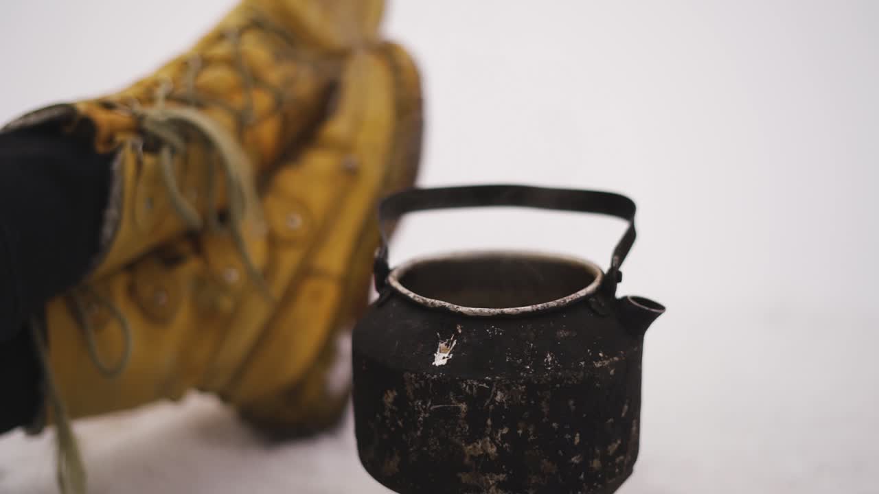 Steaming pot in the snow and hiker boots lying on the ground. Winter trip. Hot drink outdoors.