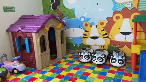 a bright indoor preschool play area with a plastic playhouse, panda ride-on cars, and vibrant animal-themed wall art. perfect for education, parenting, and childcare content. shot in 4k.