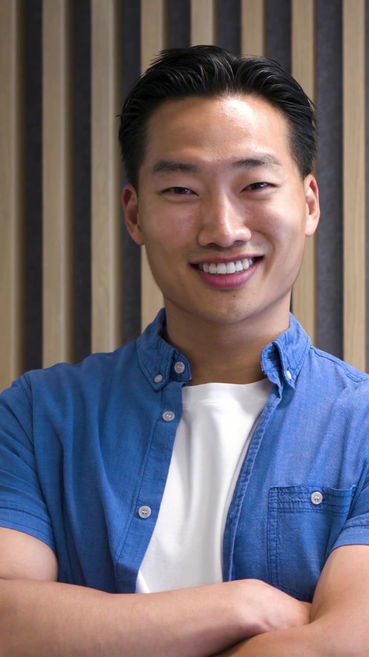 Video verticale: Uomo coreano che si dondola, sorridendo e ridendo, a braccia incrociate in studio, vicino a una parete a doghe, con una camicia blu.