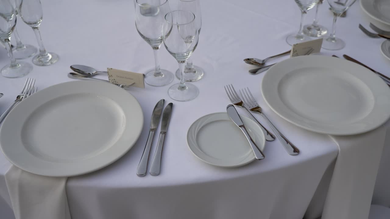 Un tavolo è stato preparato per una cena in una sala ricevimenti. Piatti, bicchieri, posate e tovaglioli sono disposti con cura. L'ambiente sembra pronto per accogliere gli ospiti e permettere loro di gustare il pasto.