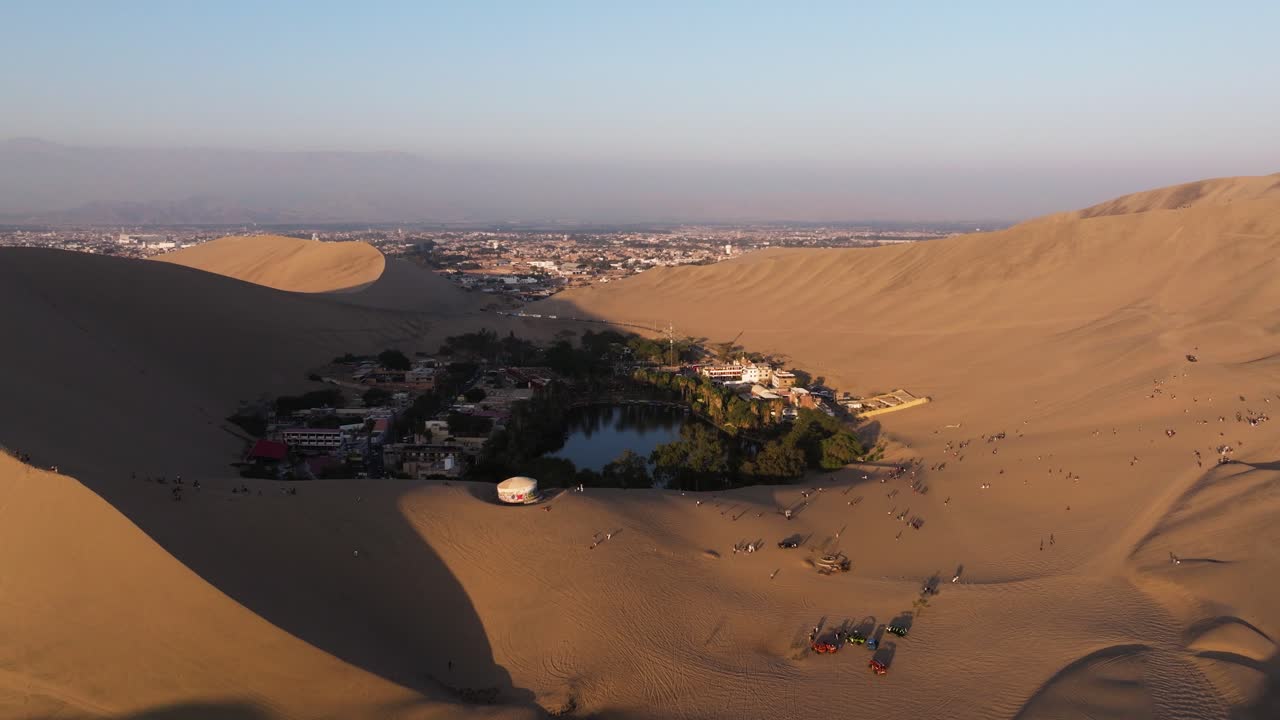 Aerial dolly establishes dune ridges framing oasis village as warm sunlight highlights sweeping sand forms