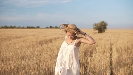 gorgeous young woman in white dress runnung in the wheat field. smiling, happy female running and holds her straw hat on her head. front view