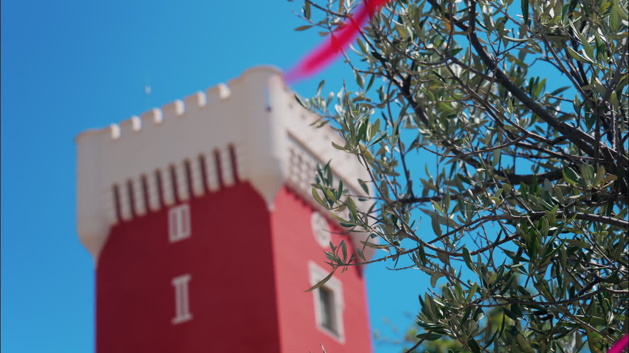 Green tree moving in the wind near the red tower of the Cremat Castle Winery over blue sky