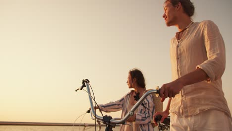 happy couple guy and girl walking together with their bicycles along the beach near the sea at sunrise. a guy in light beachwear and a girl in a blue white shirt walk with their bicycles along the beach against the sky at sunrise