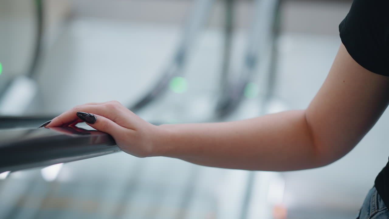 Primo piano della mano di una donna con lunghe unghie nere smaltate appoggiata al corrimano di una scala mobile durante la salita; sullo sfondo, luci bokeh verdi sfocate in un ambiente interno moderno.