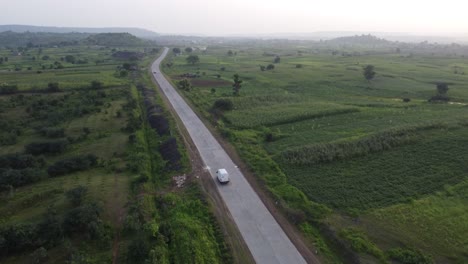 Drone-Siguiendo-Una-Furgoneta-De-Transporte-De-Mercancías-Que-Viaja-Por-Una-Carretera-India-Rural-De-Buena-Calidad-Con-Los-Alrededores-De-Tierras-De-Cultivo-Verdes-Y-Casas-De-Aldea