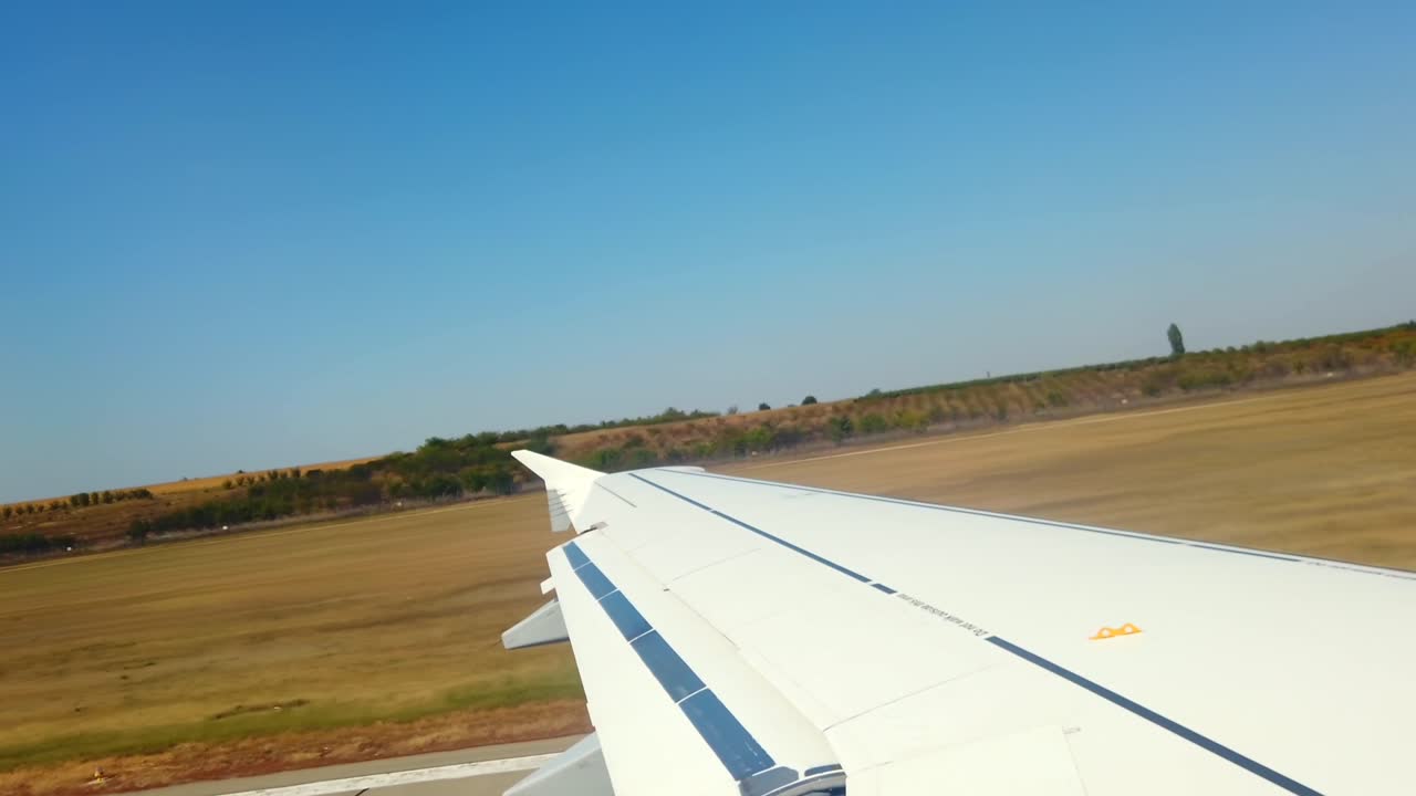 An airplane descends toward the airport, offering a wide view of sprawling fields and a clear blue sky. The landscape below showcases open land and distant trees