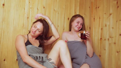 two ladies wrapped in towels relax inside wooden sauna, one adjusting her long hair with soft smile while the other makes casual hand gesture holding tea cup, sharing light calm moment