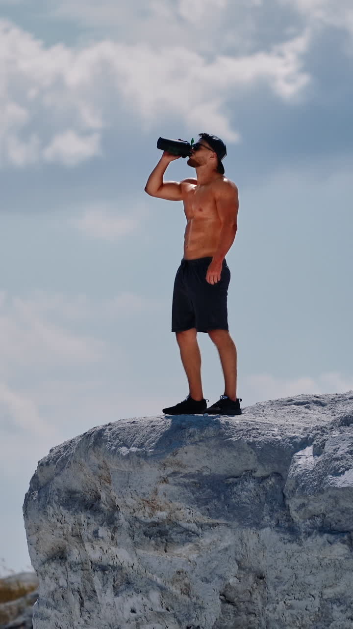 Muscular male in shorts only drinking water after hard workout outdoors. Bodybuilder standing on white rock with flask on the sky background. Vertical video