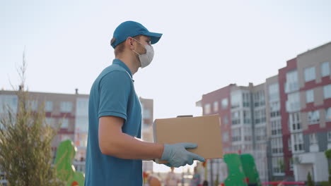 the male deliveryman in a cap and a protective mask and gloves goes with a box in his hands and carries a parcel to the customer. delivering online home orders.