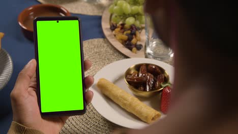 man looking at green screen mobile phone sitting around muslim family table at home eating iftar meal breaking daily fast during ramadan