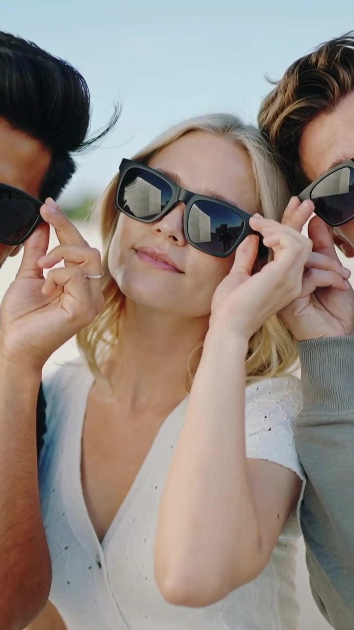 Verticale video: Zon raakt vrouw in het midden aan en twee mannen tillen zonnebrillen op op het strand, poserend, in denim