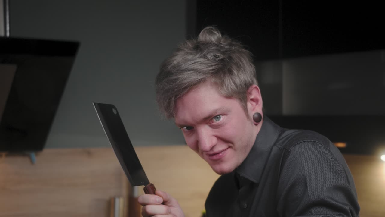 mirada malvada en la cámara y lamiendo el cuchillo de cortar con risa malvada por un joven chef psicópata loco con una elegante camisa negra con un aspecto alternativo y un tatuaje moderno, cocina de aspecto elegante a cámara lenta