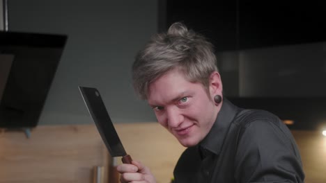 Evil-stare-in-camera-and-licking-chopping-knife-with-evil-laugh-by-young-crazy-psychopath-male-chef-in-an-elegant-black-shirt-with-an-alternative-look-and-tattoo-Modern,-fancy-looking-kitchen-Slow-Mo