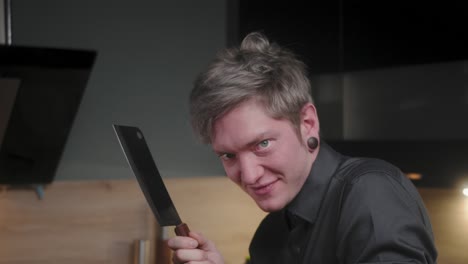 mirada malvada en la cámara y lamiendo el cuchillo de cortar con risa malvada por un joven chef psicópata loco con una elegante camisa negra con un aspecto alternativo y un tatuaje moderno, cocina de aspecto elegante a cámara lenta