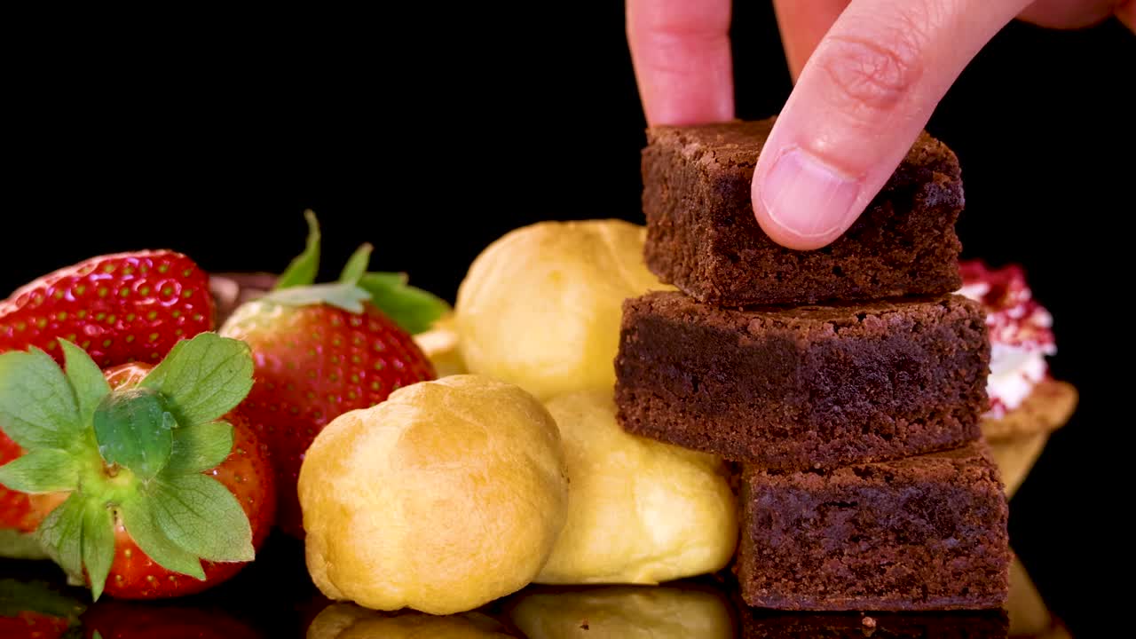 A hand picks up a chocolate brownie from a neatly arranged selection of desserts, including strawberries and pastries, on a reflective black surface under bright studio lighting