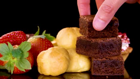 a hand picks up a chocolate brownie from a neatly arranged selection of desserts, including strawberries and pastries, on a reflective black surface under bright studio lighting