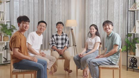 asian teen group smiling to camera while sitting in chairs forming a circle discussing at home