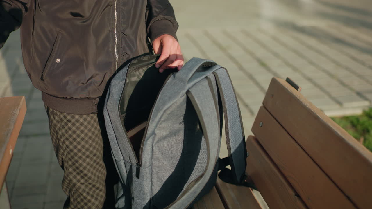 Close up of student packing book into backpack while standing beside wooden bench on paved outdoor area with natural sunlight creating strong shadows on ground