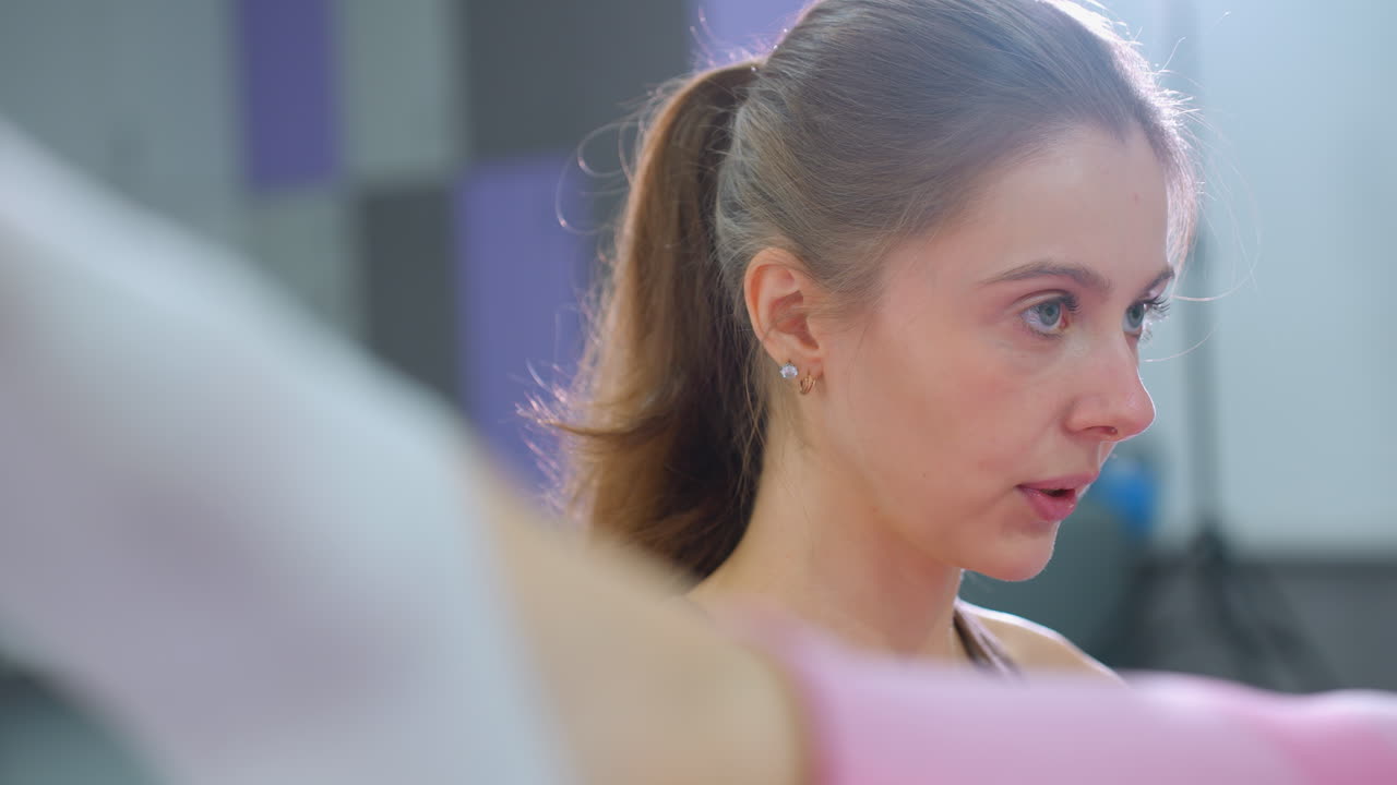 핑크색 운동복을 입고 운동 중 다리 움직임의 클로즈업, 갈색을 입은 여성이 근처에 초점을 맞추고, 자연 빛에 의해 백라이트, 흐릿한 체육관 배경