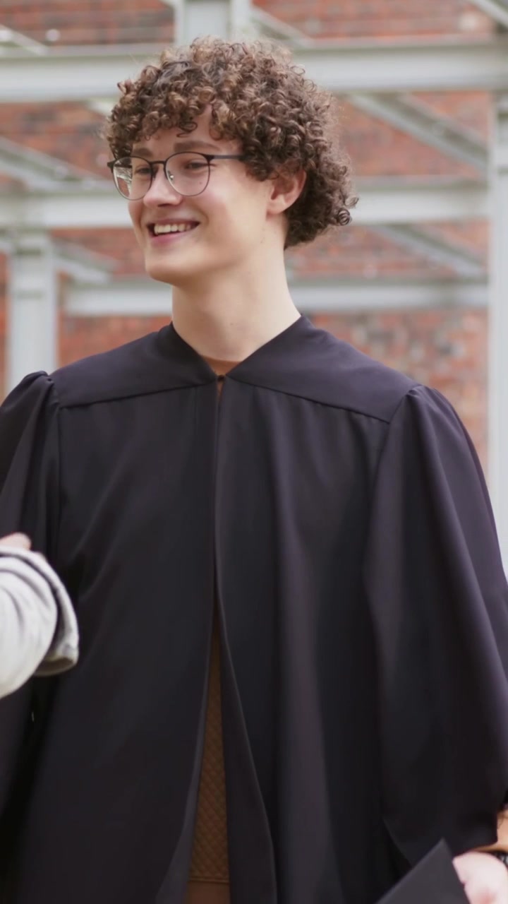 Video verticale: Laureato in toga che festeggia, batte il cinque a un collega che si avvicina da sinistra nel cortile universitario, tenendo il tocco.