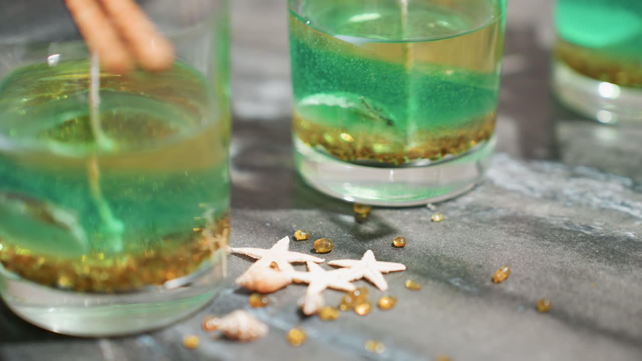 Fair skinned lady gently drops decorative beads and starfish-shaped embellishments beside glass jars filled with colorful wax mixture during home candle-making process on smooth kitchen countertop