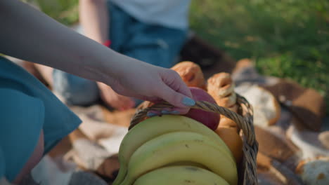 un primer plano de una mujer colocando una canasta llena de plátanos, manzanas y otras frutas en una manta de picnic a cuadros. se ve a un niño borroso sentado en la manta en un campo de hierba